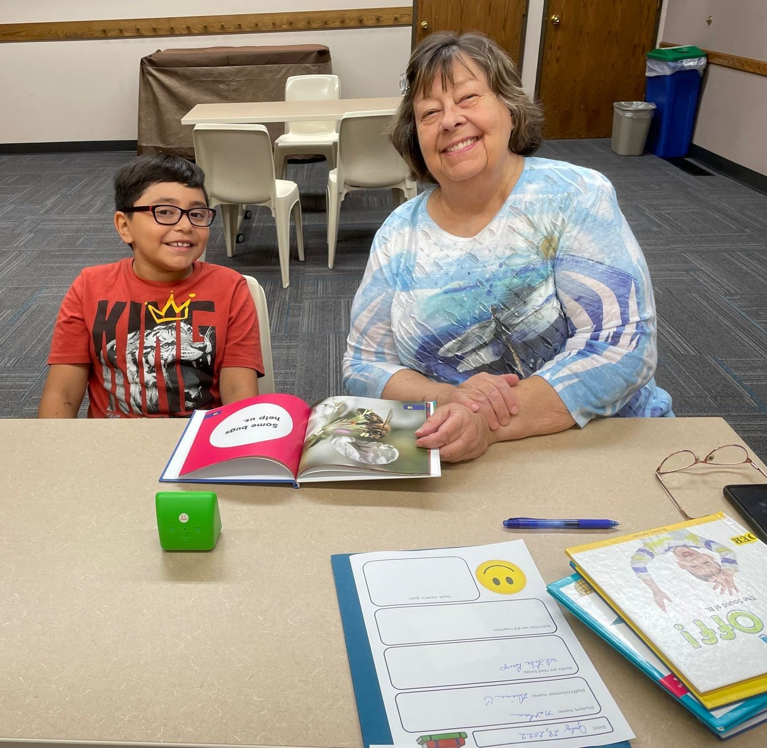 An older woman sits at a table with a young boy and they are reading a book together